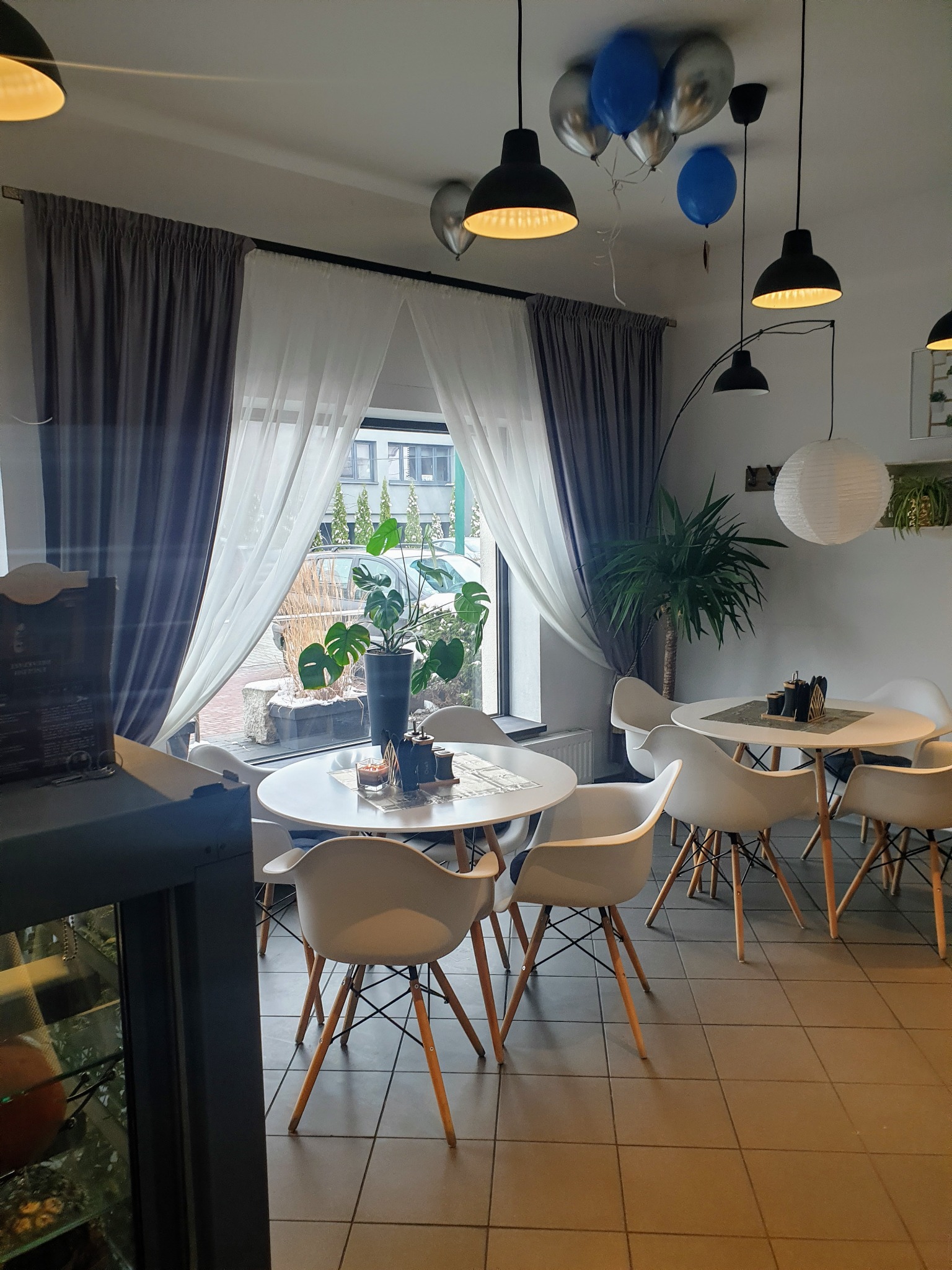 Wnętrze kawiarni z białymi krzesłami i okrągłymi stolikami, zasłonięte szarymi i białymi zasłonami, z dekoracjami w postaci balonów i roślin.