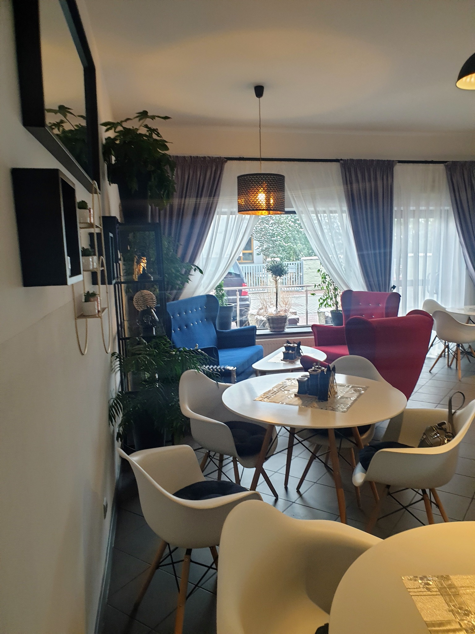 Wnętrze kawiarni z dużym oknem, przez które widać drzewa, z zasłonami w odcieniach szarości i bieli, z okrągłymi stolikami i krzesłami w stylu skandynawskim, z roślinami doniczkowymi i lampą...