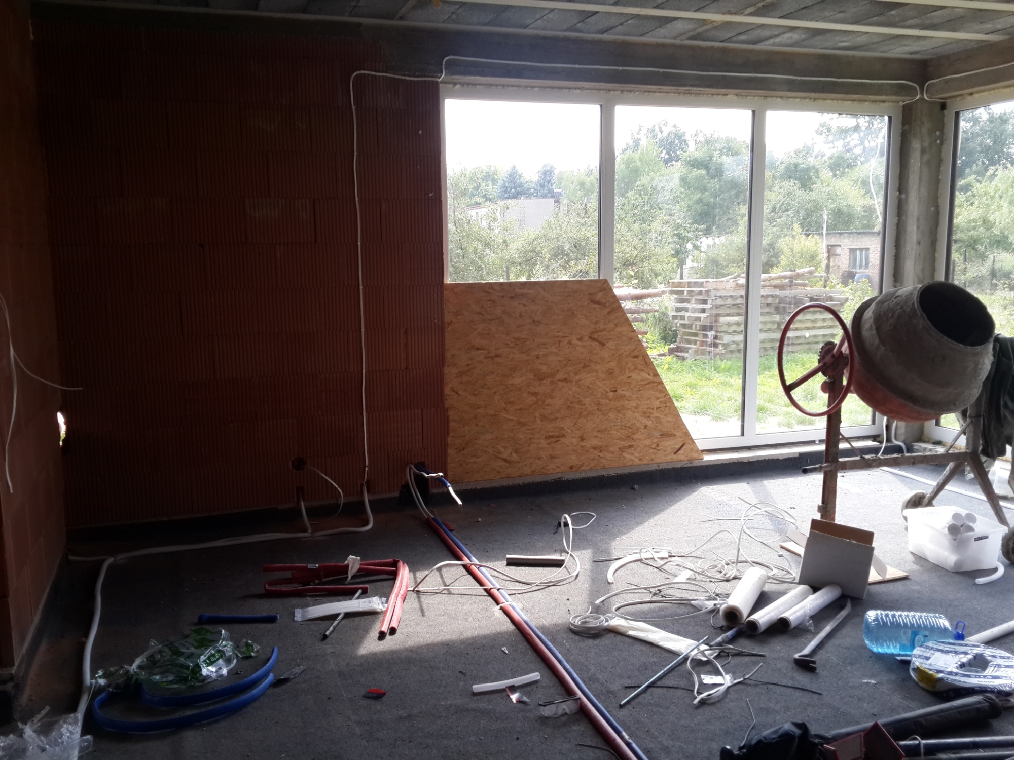 Wnętrze w trakcie prac wykończeniowych: ściany z czerwonej cegły, okno z widokiem na zieleń, instalacje elektryczne i hydrauliczne, płyta OSB oparta o okno, betoniarka i narzędzia na podłodze.