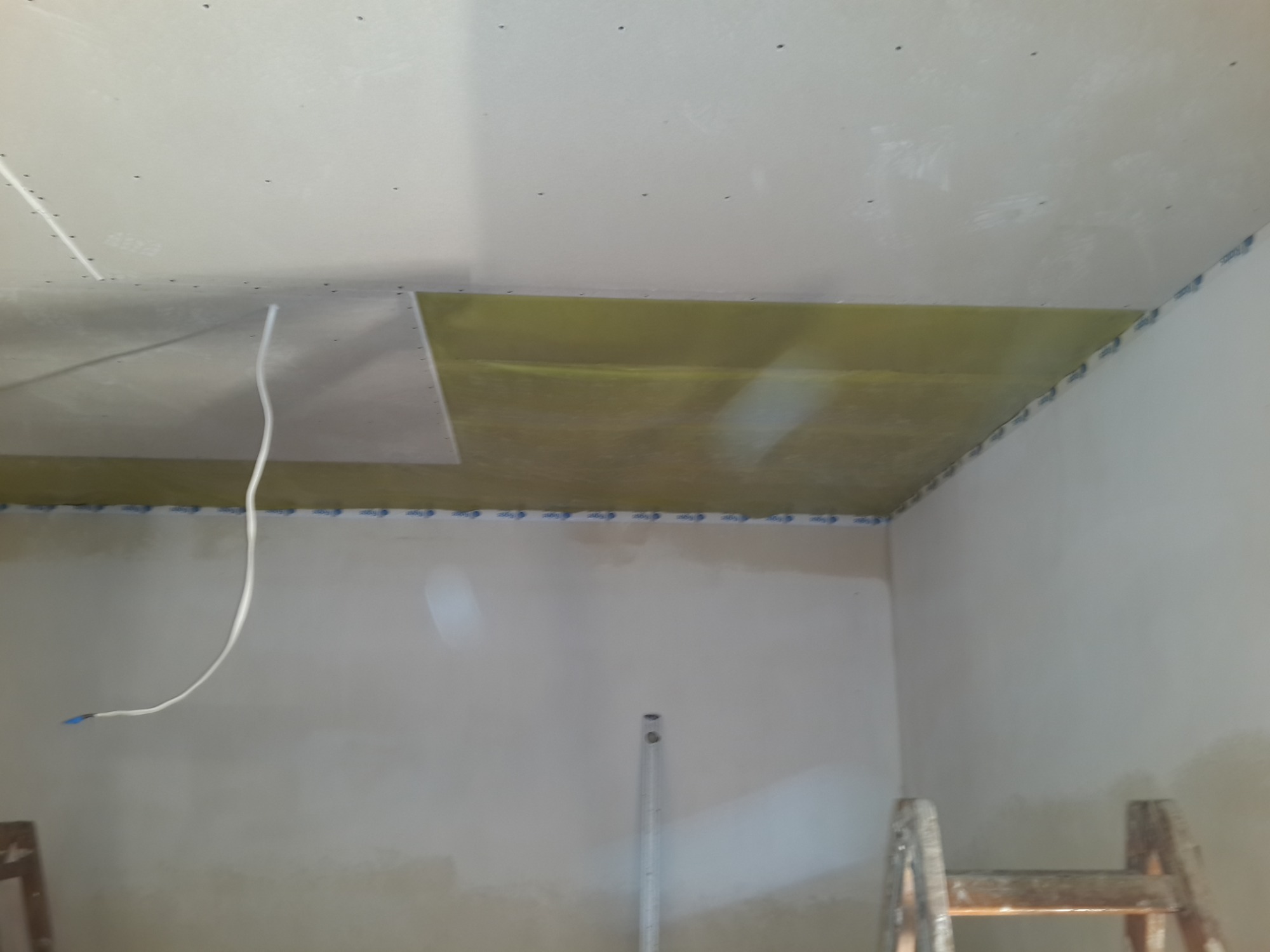 Montaż płyty gipsowo-kartonowej na suficie z widoczną izolacją, taśmą maskującą na krawędziach i kablem zwisającym ze ściany, w tle drabina i poziomica.