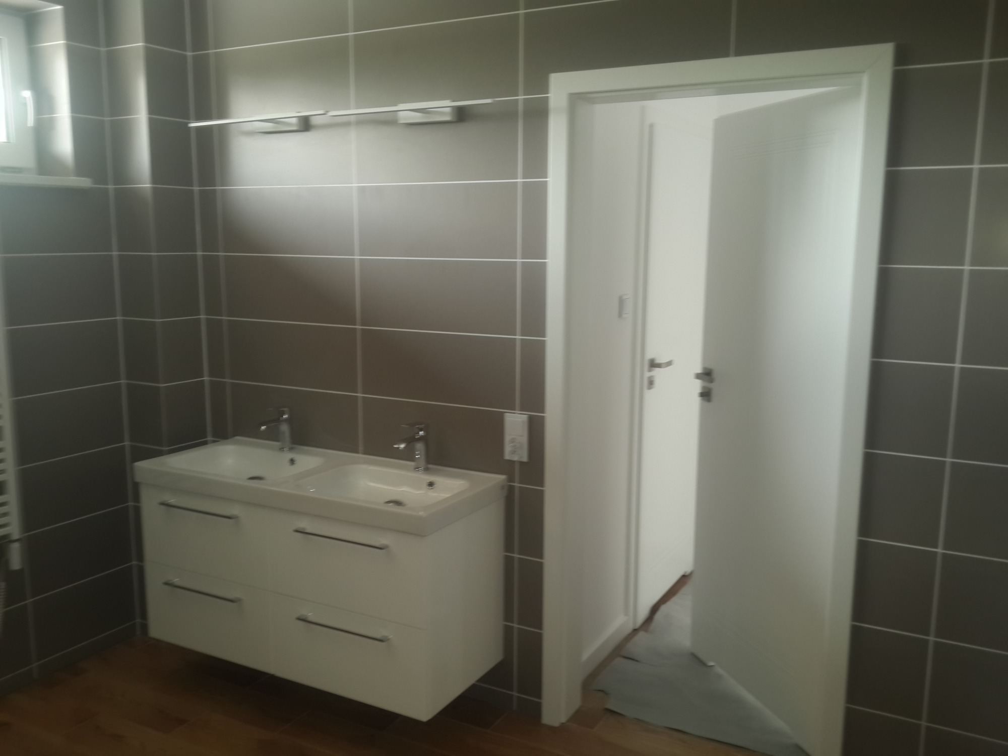 Nowoczesna łazienka z szarymi, kwadratowymi płytkami, białą szafką pod umywalką z dwiema umywalkami i uchwytami oraz otwartymi, białymi drzwiami.