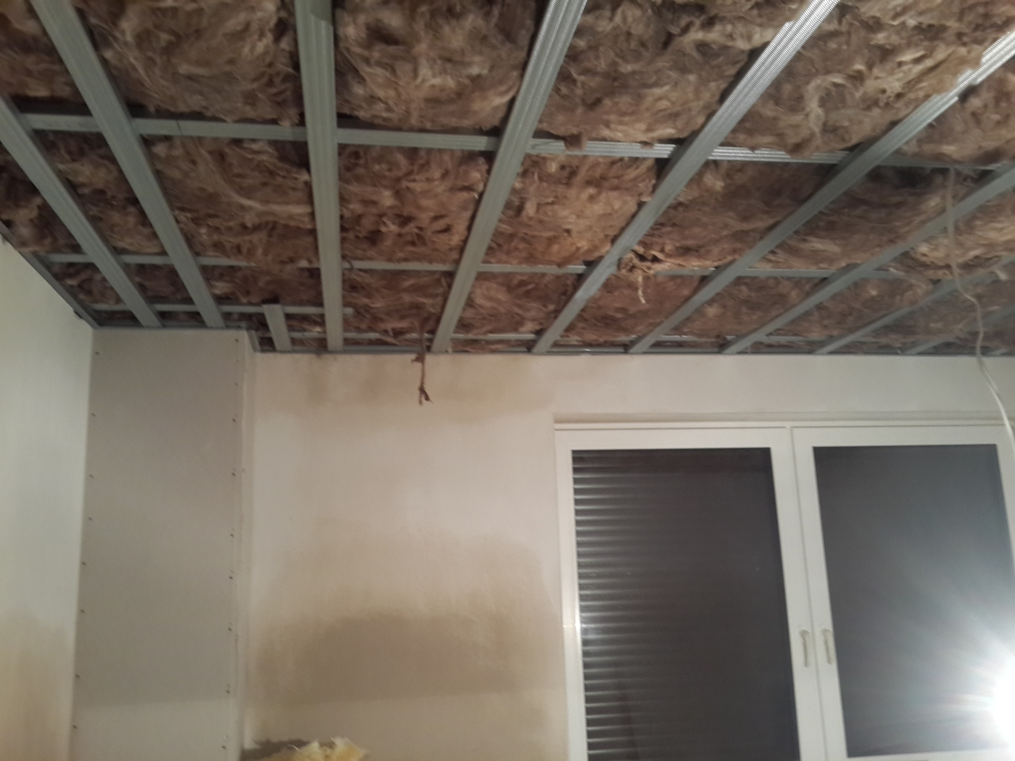 Montaż sufitu podwieszanego z izolacją z wełny mineralnej, widoczne metalowe profile i częściowo wykończona ściana z oknem.