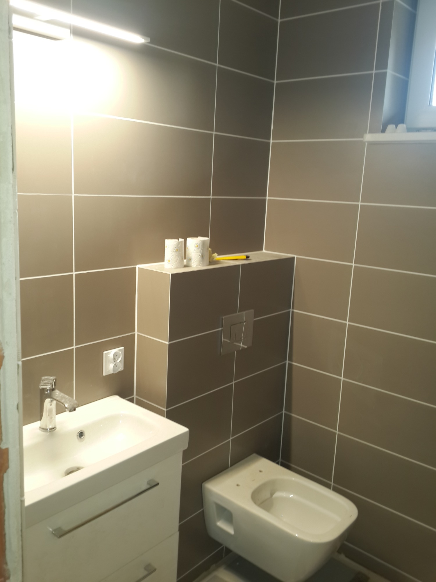 Nowoczesna łazienka z brązowymi płytkami, białą umywalką z szafką, podwieszaną toaletą i rolkami papieru toaletowego na zabudowie stelaża. Widoczny fragment niedokończonego wykończenia przy...