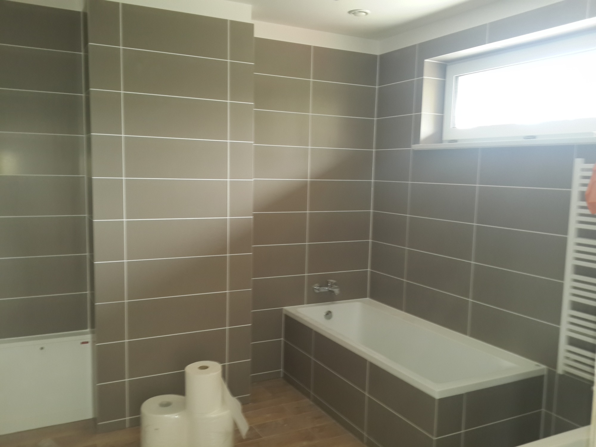 Wykończona łazienka z brązowymi, poziomymi płytkami, białą wanną i grzejnikiem drabinkowym; na podłodze leżą rolki papieru toaletowego.