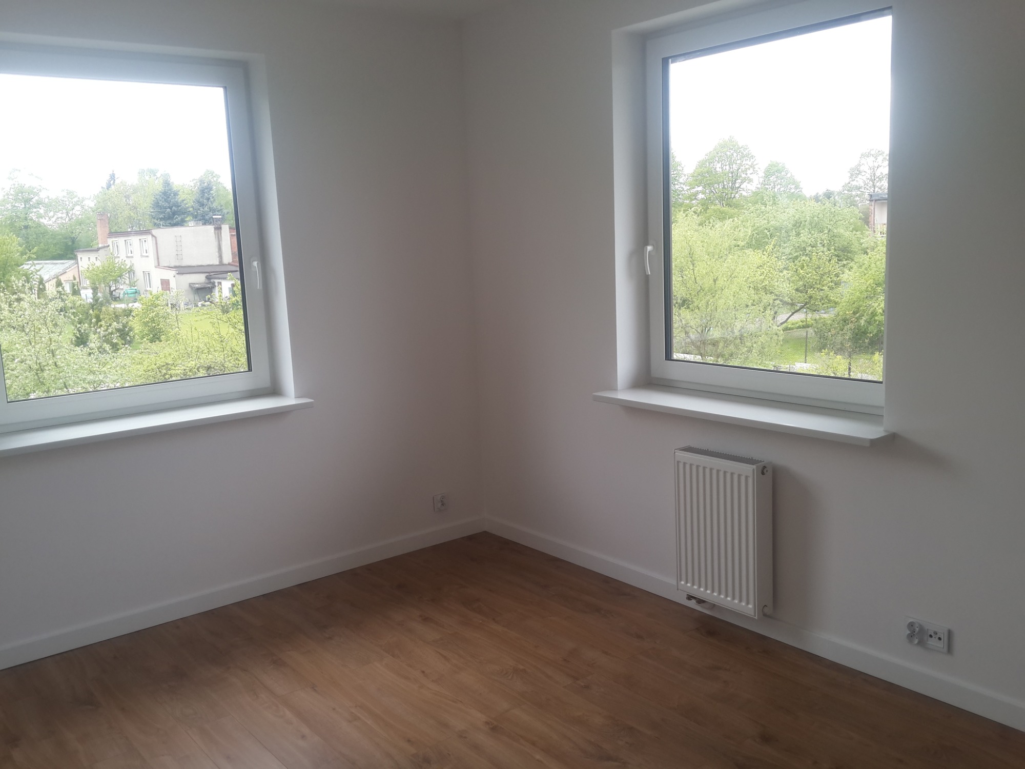 Pusty pokój po remoncie w Łodzi z drewnopodobną podłogą, białymi ścianami, dwoma oknami z widokiem na zieleń i grzejnikiem pod jednym z nich.