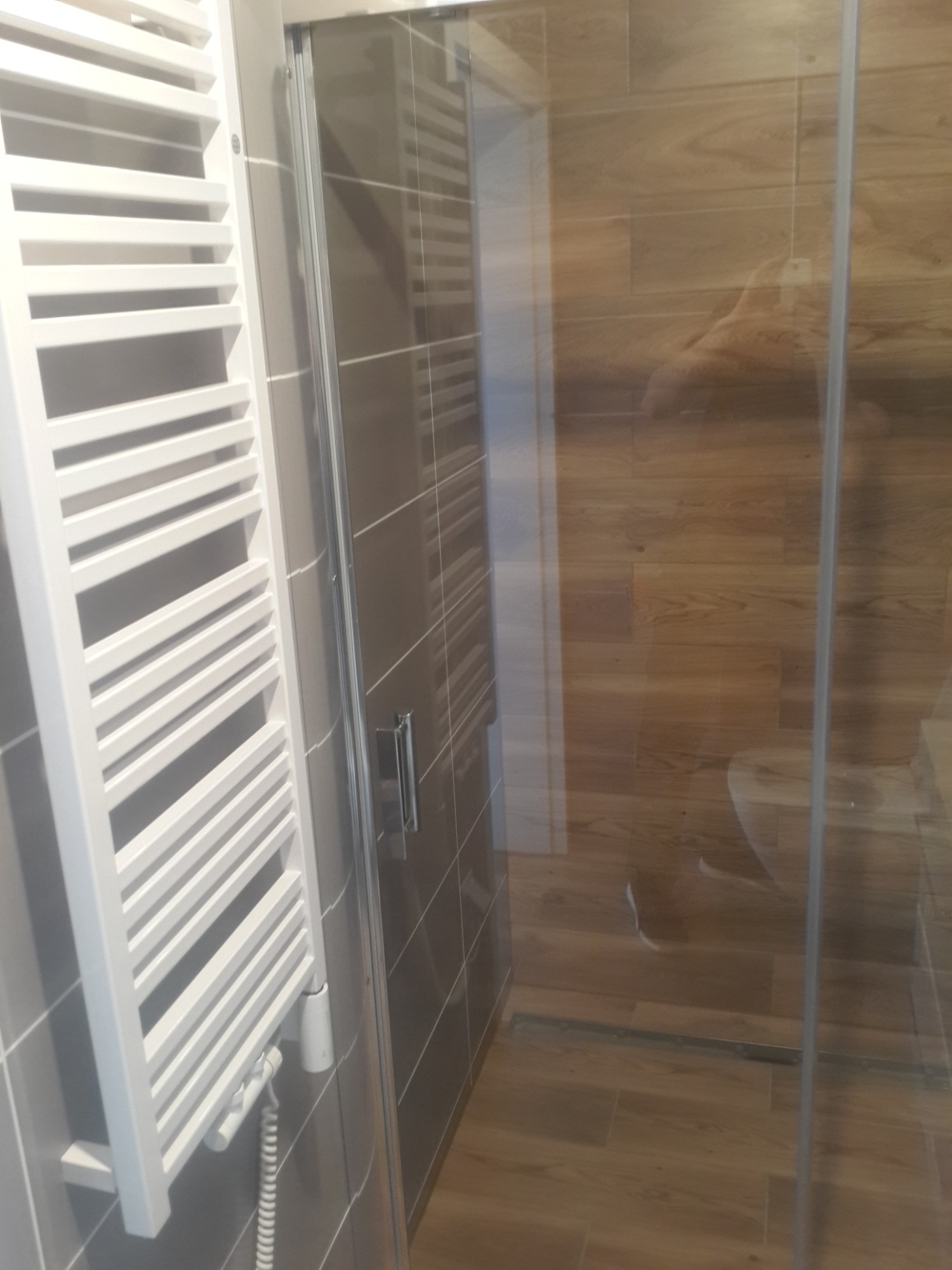 Nowoczesna łazienka z grzejnikiem drabinkowym, szarą glazurą i kabiną prysznicową z imitacją drewna na ścianach.