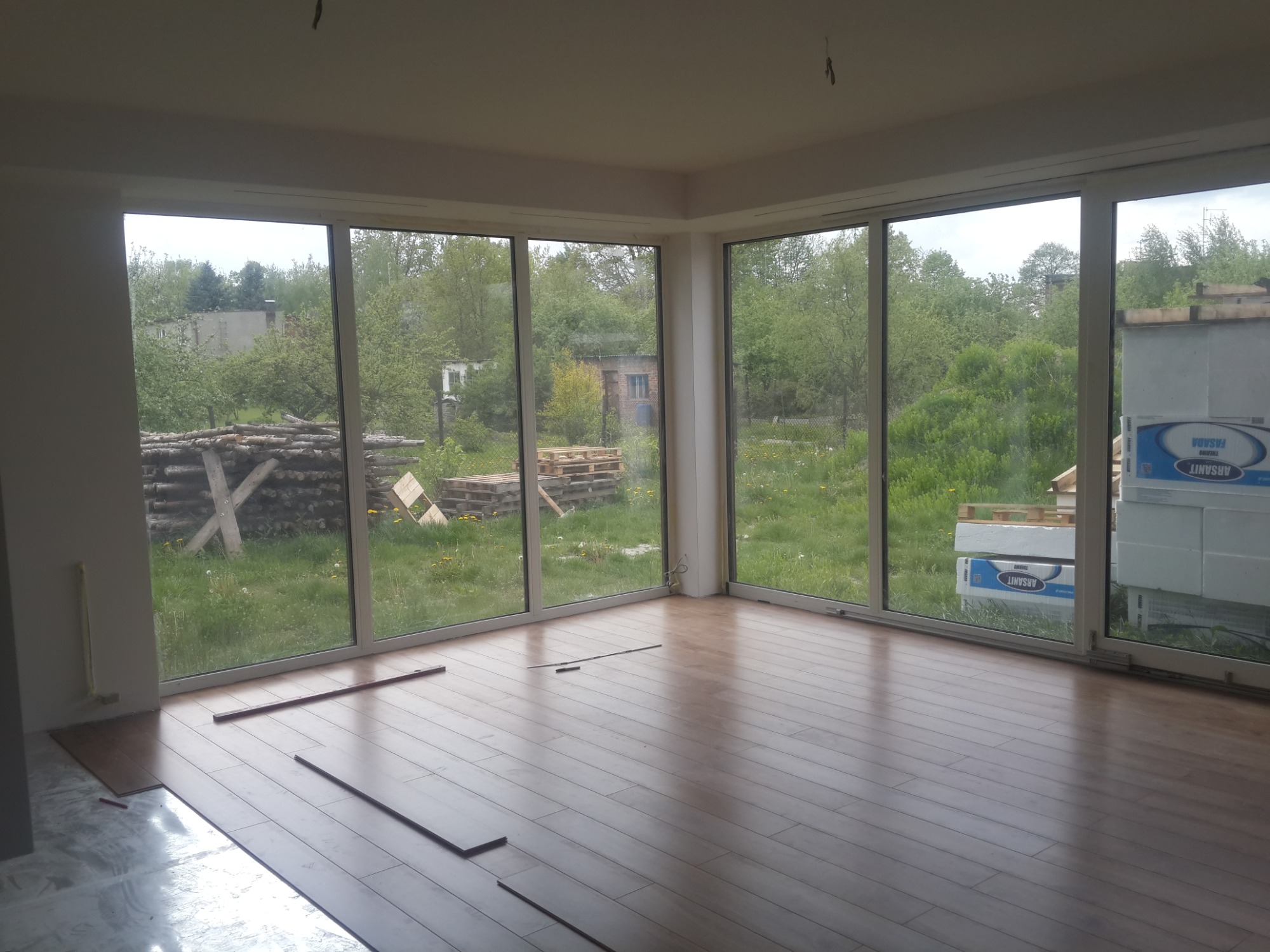 Wnętrze w trakcie układania drewnianej podłogi, widoczne duże okna z widokiem na zewnątrz, palety i drewno za oknem.