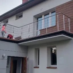 Dwupiętrowy dom z balkonem zabezpieczonym nowoczesną, poziomą konstrukcją ze stali nierdzewnej. Dwie osoby na balkonie, jedna w różowej bluzie, druga w czerwonej kurtce. Wykończenie balkonu drewnem.