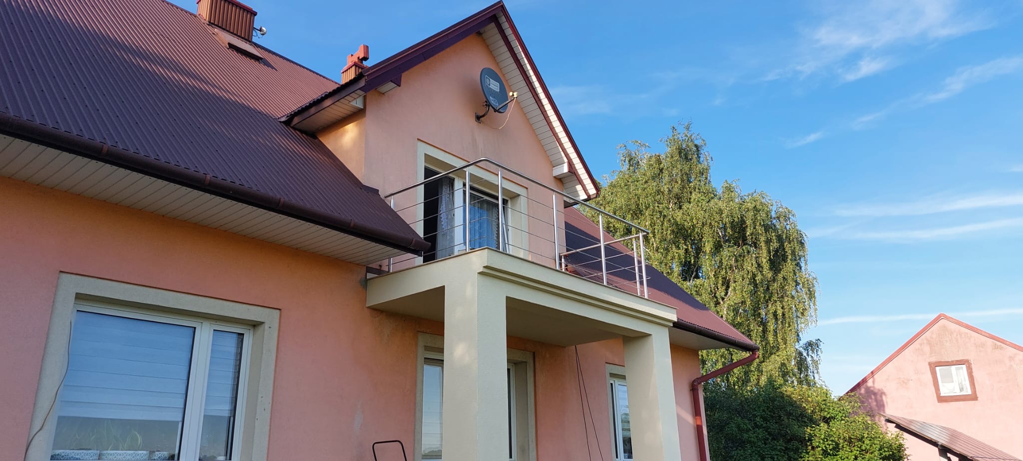 Dom z balkonem zabezpieczonym nowoczesną, minimalistyczną balustradą ze stali nierdzewnej, widoczną na tle błękitnego nieba i zielonego drzewa.