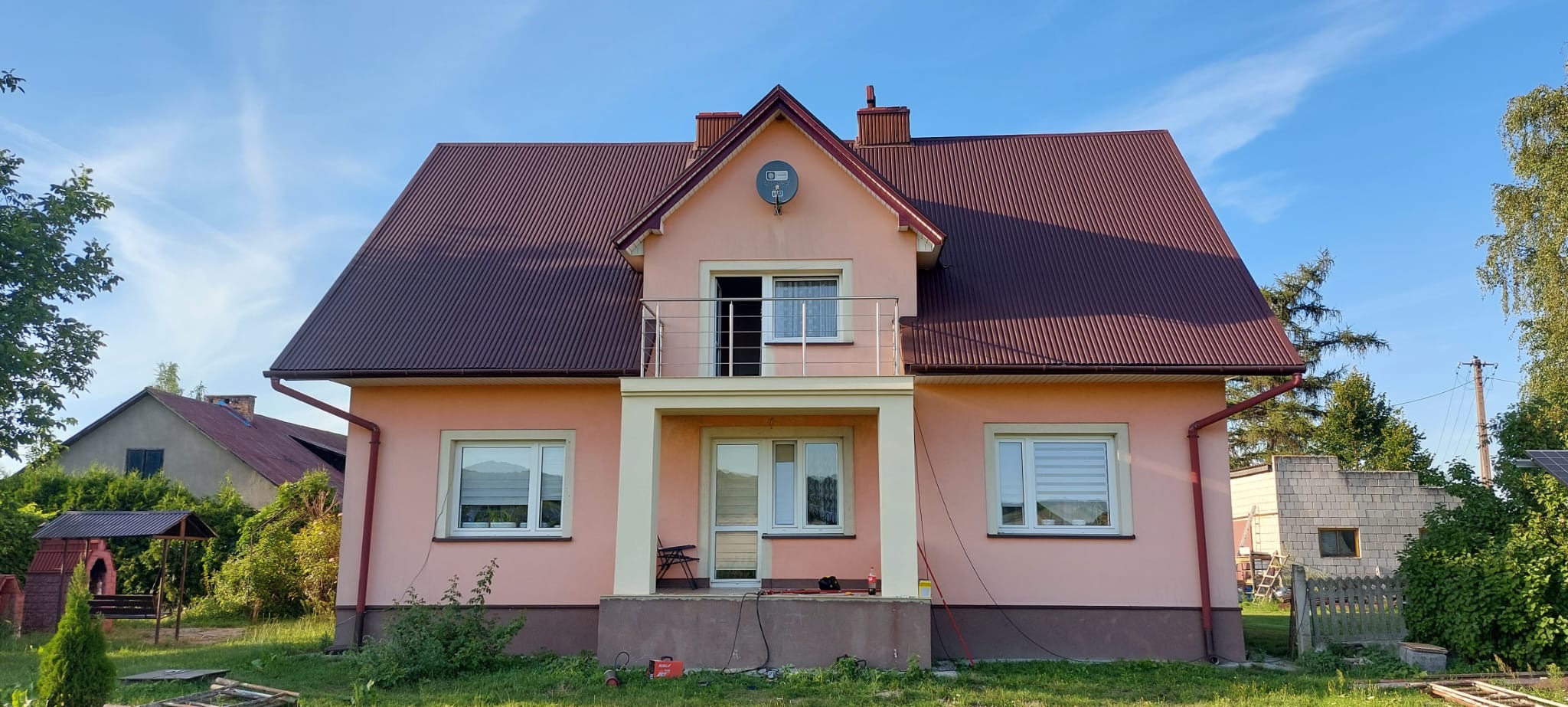 Różowy dom jednorodzinny z balkonem zabezpieczonym metalową barierką, widok z zewnątrz w słoneczny dzień.