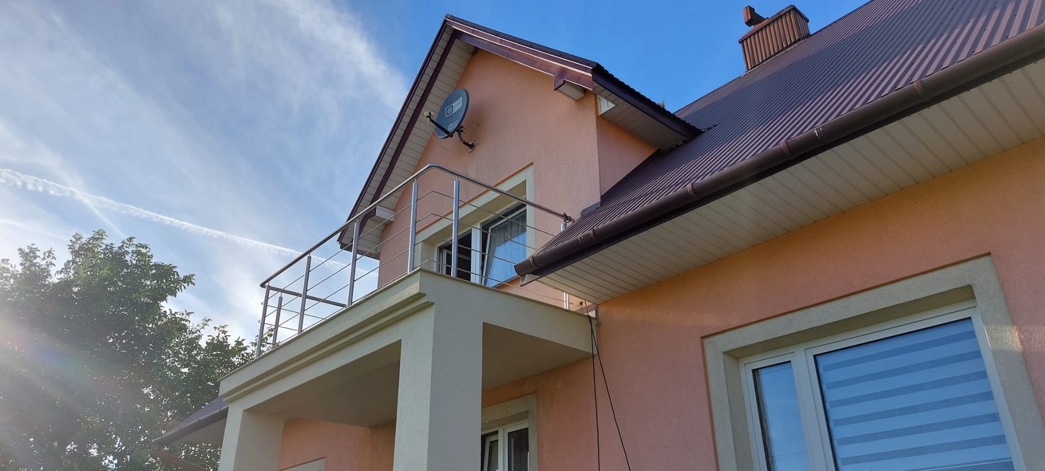 Nowoczesna, stalowa balustrada balkonowa z wypełnieniem linkowym, zamontowana w domu o różowej elewacji i brązowym dachu, widoczna antena satelitarna.