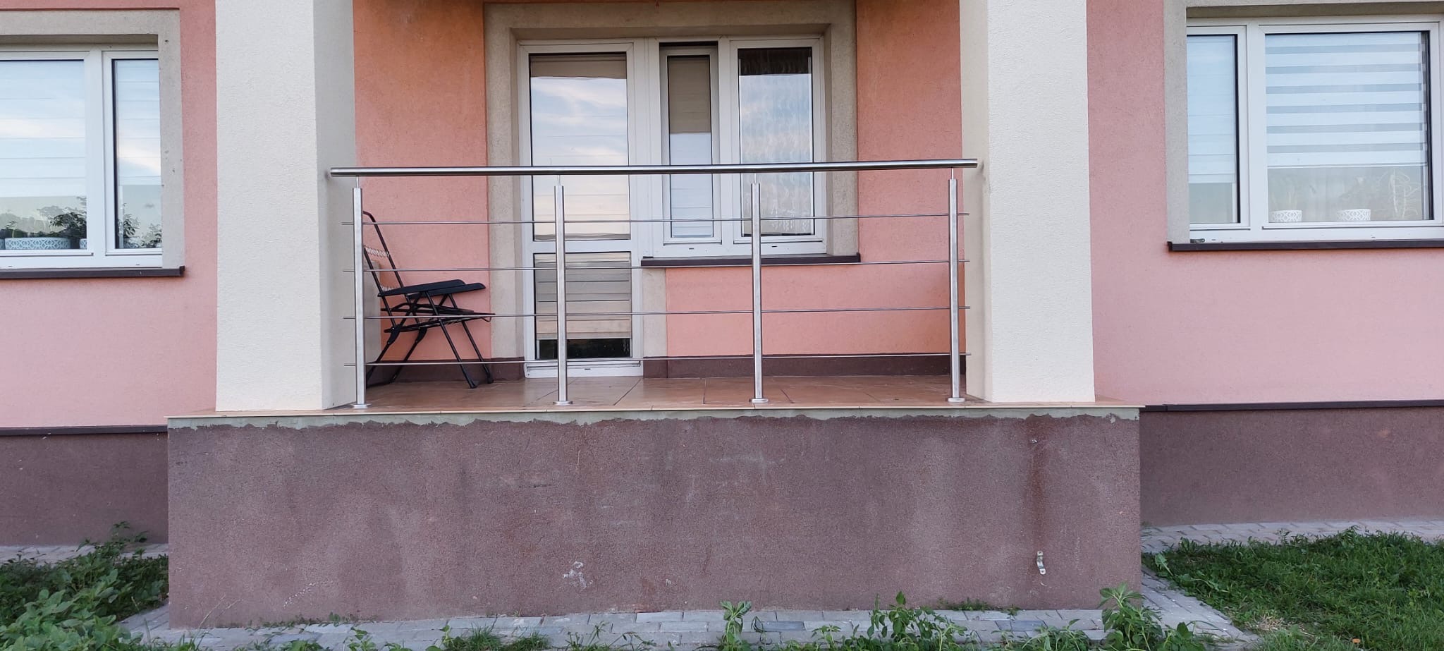 Balkon z poziomą, metalową konstrukcją zabezpieczającą, widziany z poziomu gruntu, z krzesłem ogrodowym w tle i oknami z roletami w tle.