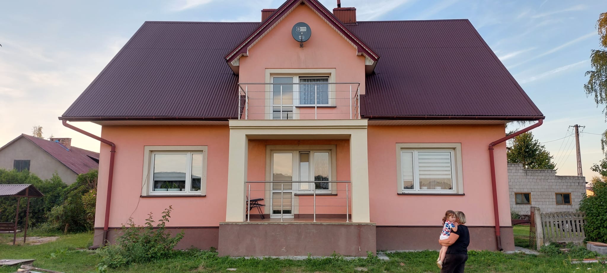 Dwukondygnacyjny dom z różową elewacją i brązowym dachem, z balkonem z metalową balustradą. W kadrze widoczna kobieta trzymająca dziecko.