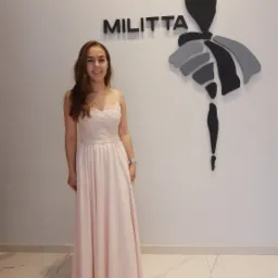 Uśmiechnięta młoda kobieta w długiej, pudrowo-różowej sukni wieczorowej z koronkową górą, stojąca na tle białej ściany z logo firmy odzieżowej.