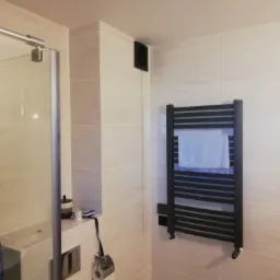 Nowoczesna łazienka z kabiną prysznicową, grzejnikiem drabinkowym z ręcznikiem, wentylatorem i ceramicznymi płytkami ściennymi w odcieniach beżu i bieli.