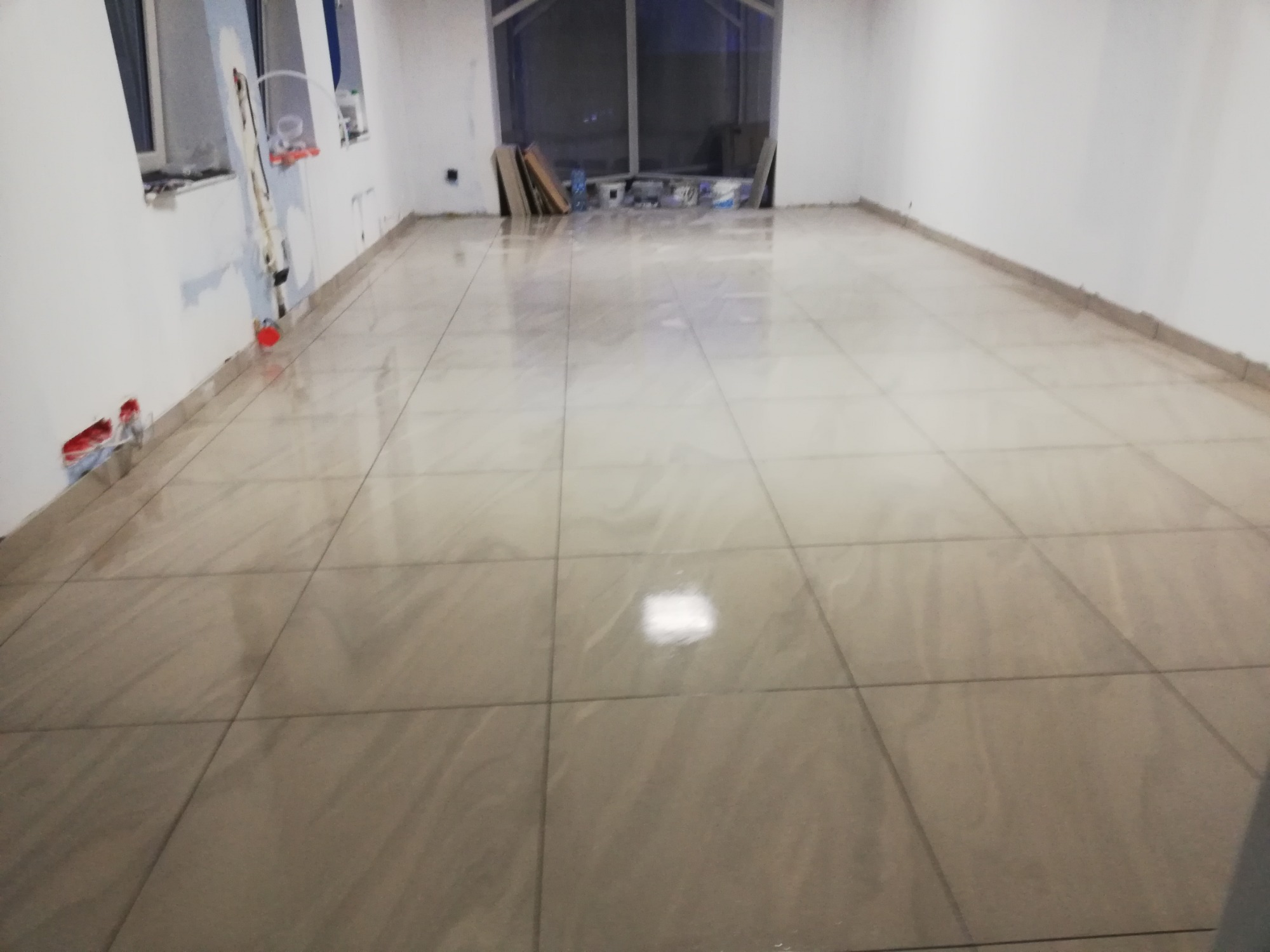 Ułożona podłoga z płytek ceramicznych w dużym, pustym pokoju, widoczne niedokończone prace przy ścianach i okablowaniu.