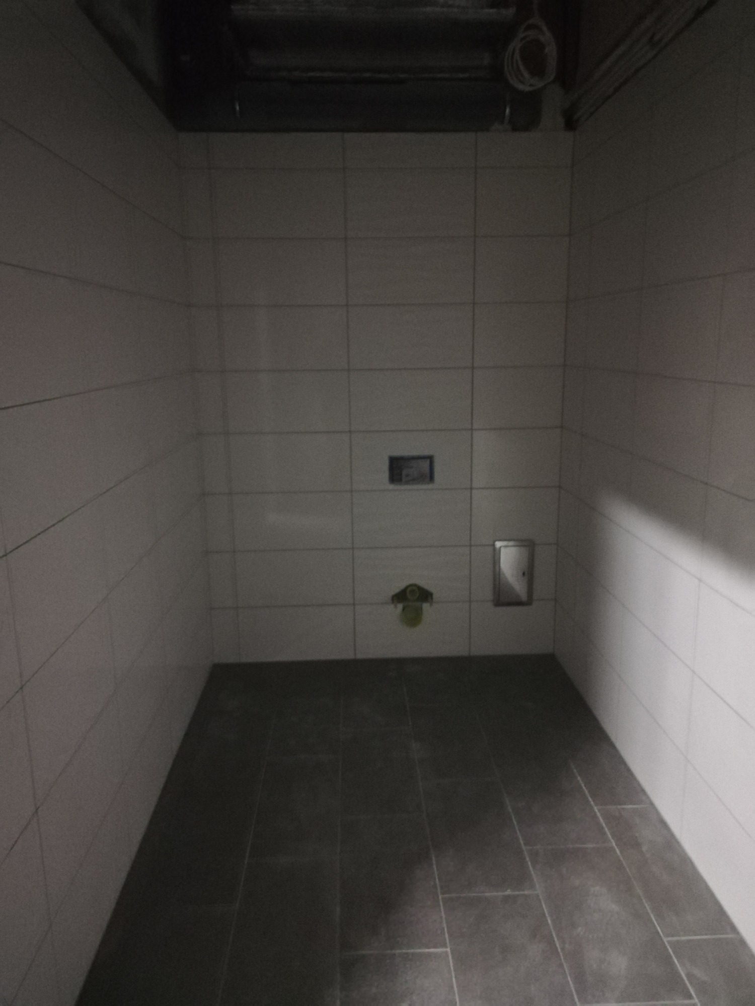 Surowy stan łazienki z ułożonymi ciemnymi płytkami podłogowymi i białymi płytkami ściennymi, widoczne instalacje pod sufitem i dwa otwory inspekcyjne na ścianie.