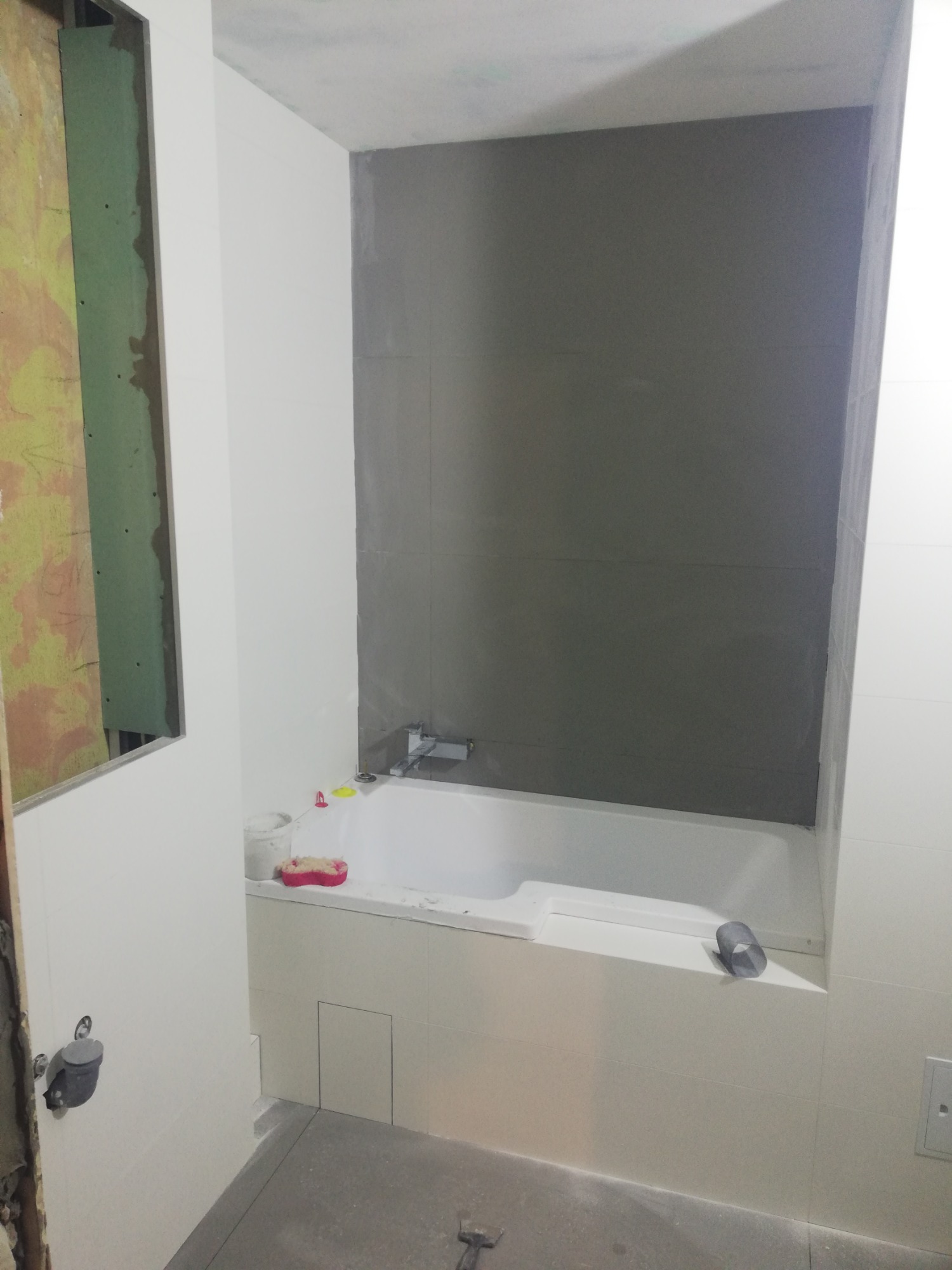 Wnęka z wanną w trakcie prac wykończeniowych, ściany częściowo pokryte szarymi płytkami, widoczne narzędzia i akcesoria łazienkowe, surowe wykończenie ściany po lewej stronie.