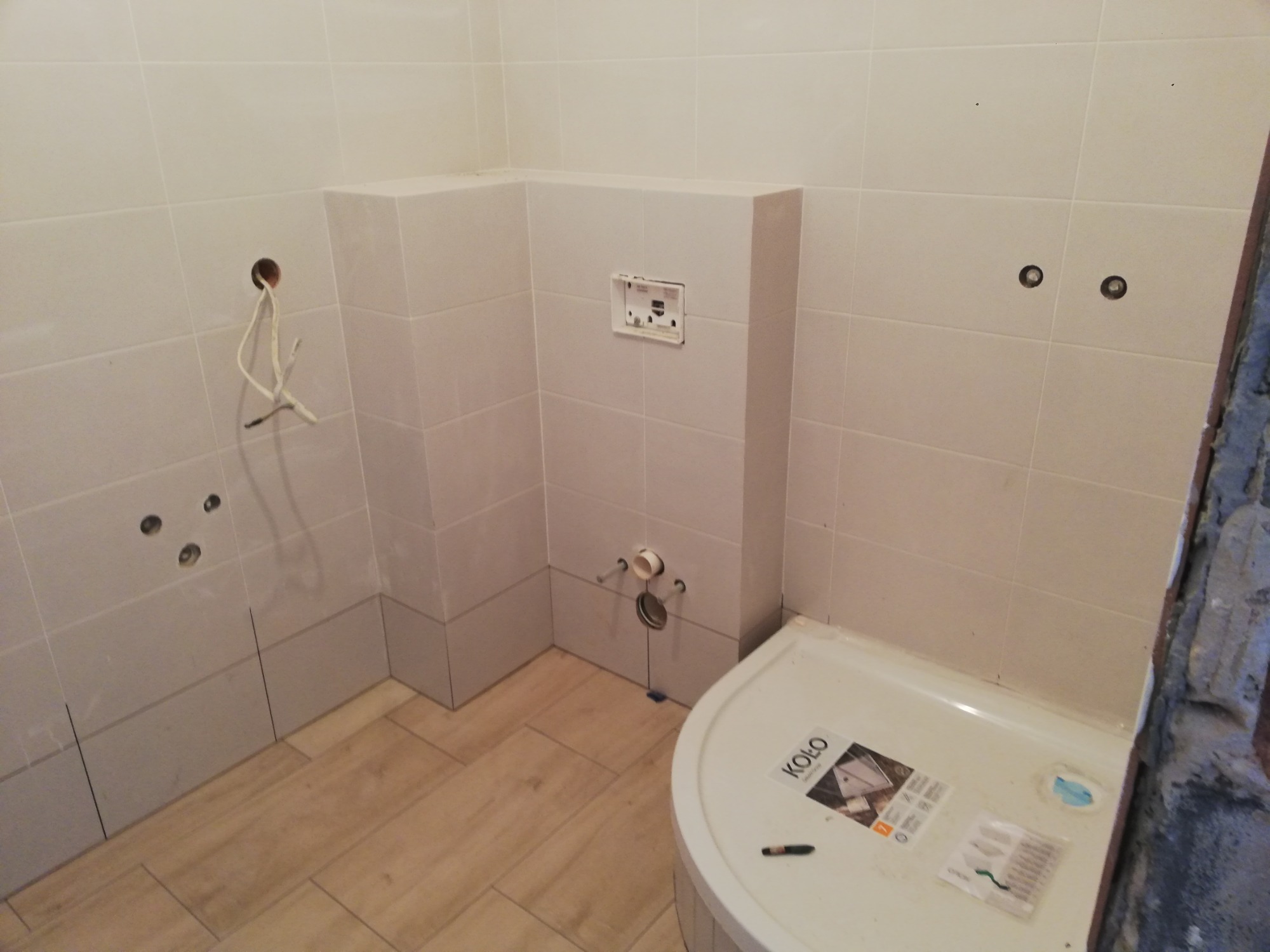 Łazienka w trakcie prac wykończeniowych: ułożone jasne płytki na ścianach i podłodze imitujące drewno, widoczne otwory na armaturę, wystające kable, częściowo zamontowany stelaż podtynkowy WC...