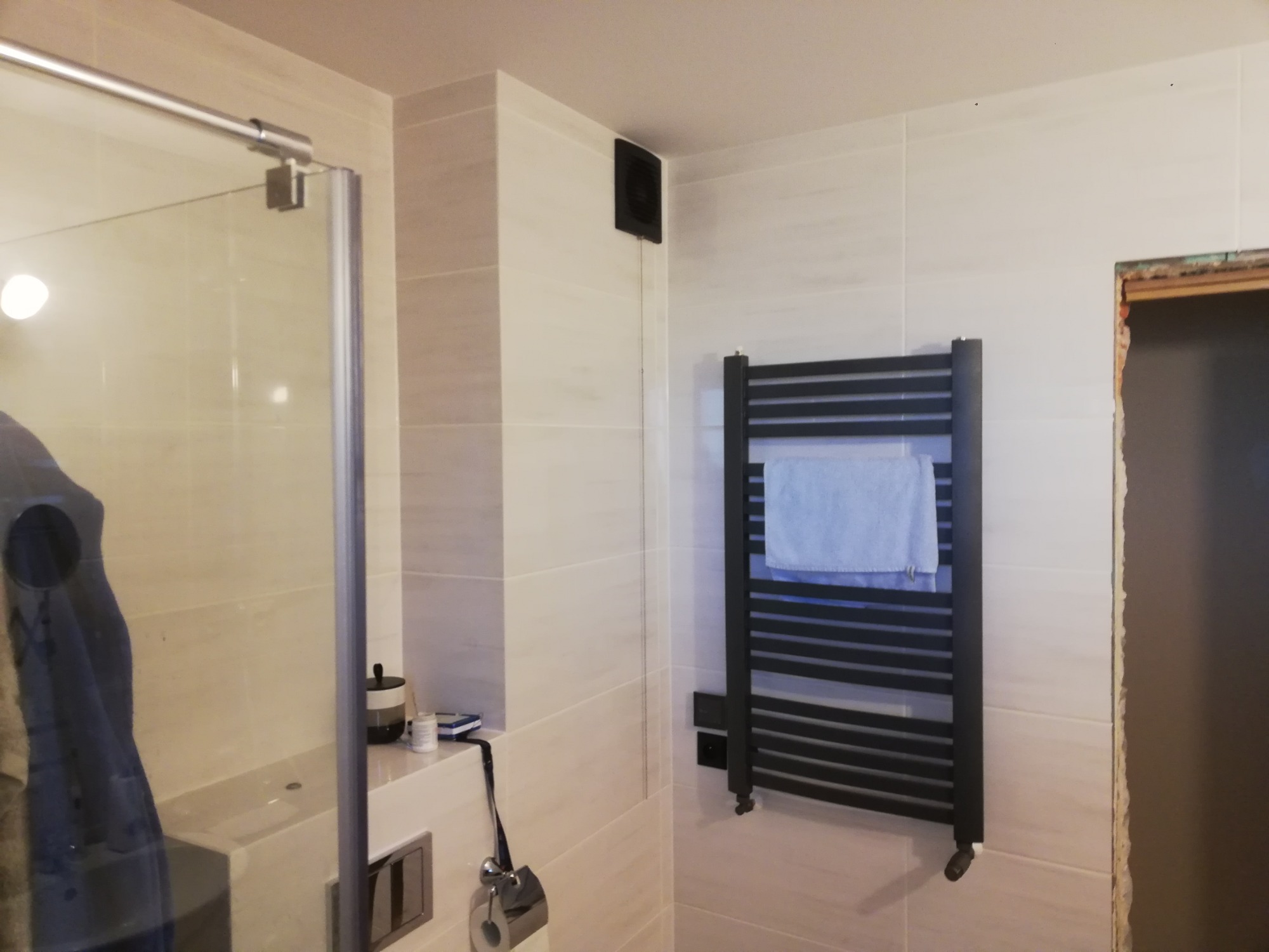 Nowoczesna łazienka z kabiną prysznicową, grzejnikiem drabinkowym z ręcznikiem, wentylatorem i ceramicznymi płytkami ściennymi w odcieniach beżu i bieli.