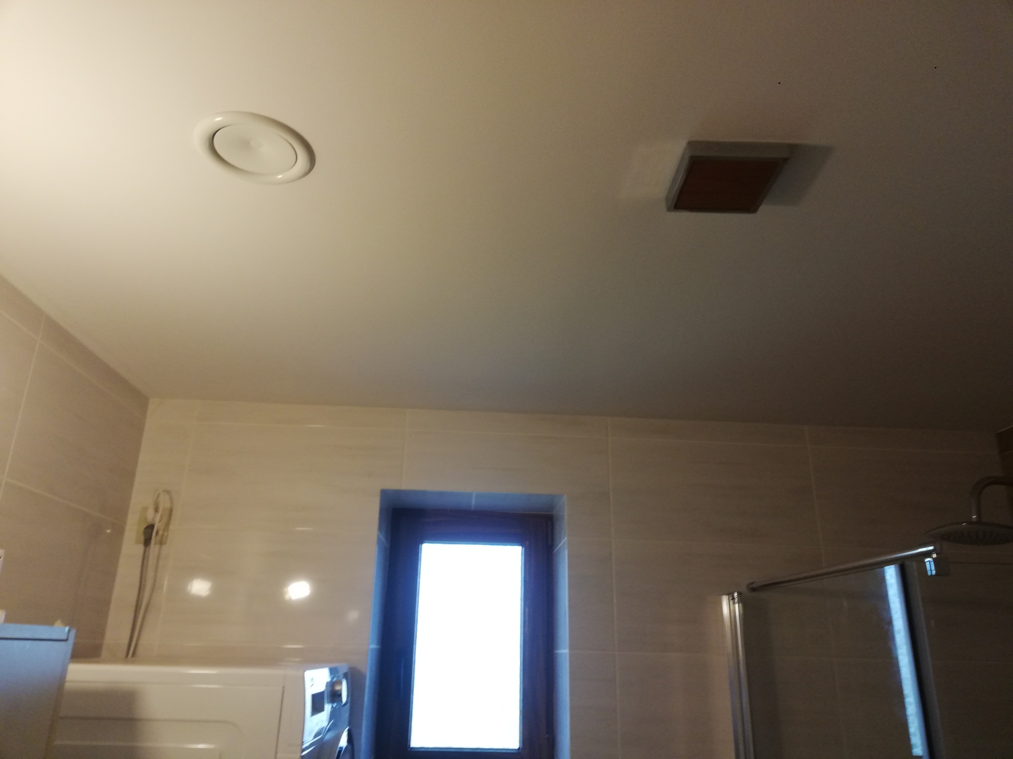 Jasna łazienka z beżowymi płytkami, widoczny fragment kabiny prysznicowej, okno i pralka, na suficie okrągły nawiewnik i kwadratowa lampa.