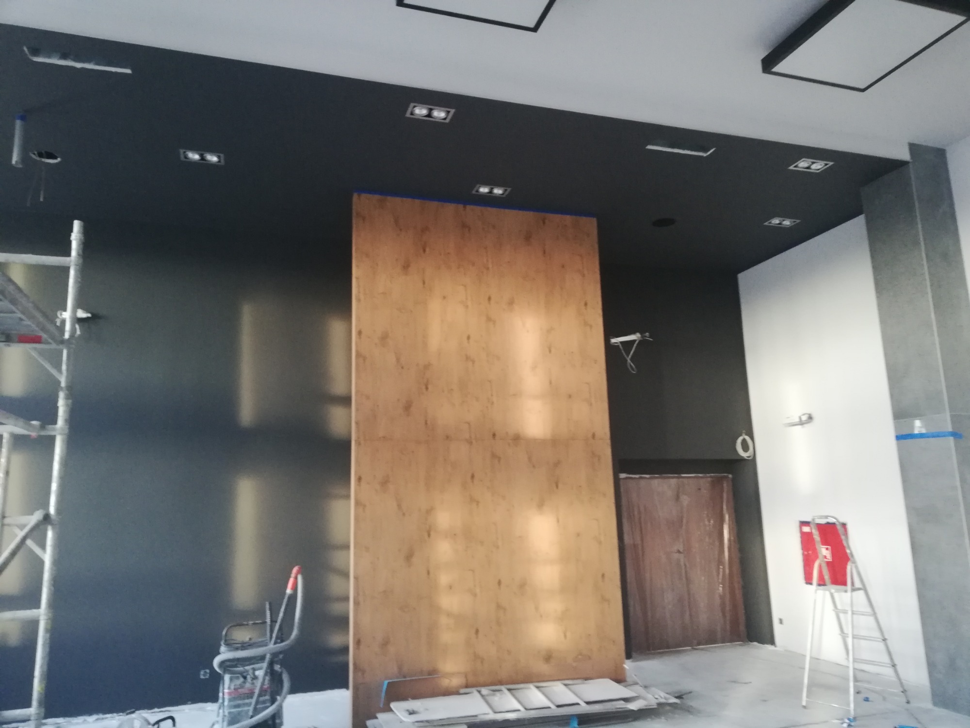 Wnętrze w trakcie prac wykończeniowych: pomalowany na czarno sufit z oświetleniem punktowym, zabezpieczona płyta okładzinowa, drabina i rusztowanie w tle.
