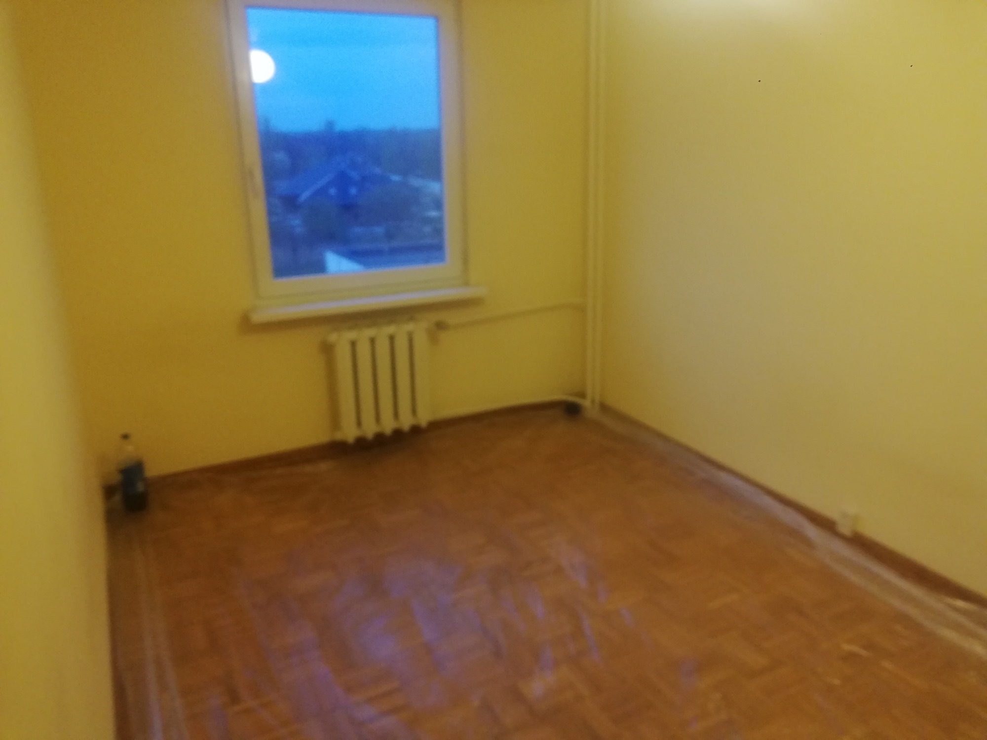 Pusty pokój z żółtymi ścianami przygotowany do prac wykończeniowych, podłoga zabezpieczona folią, widoczny grzejnik pod oknem z widokiem na budynki w oddali.
