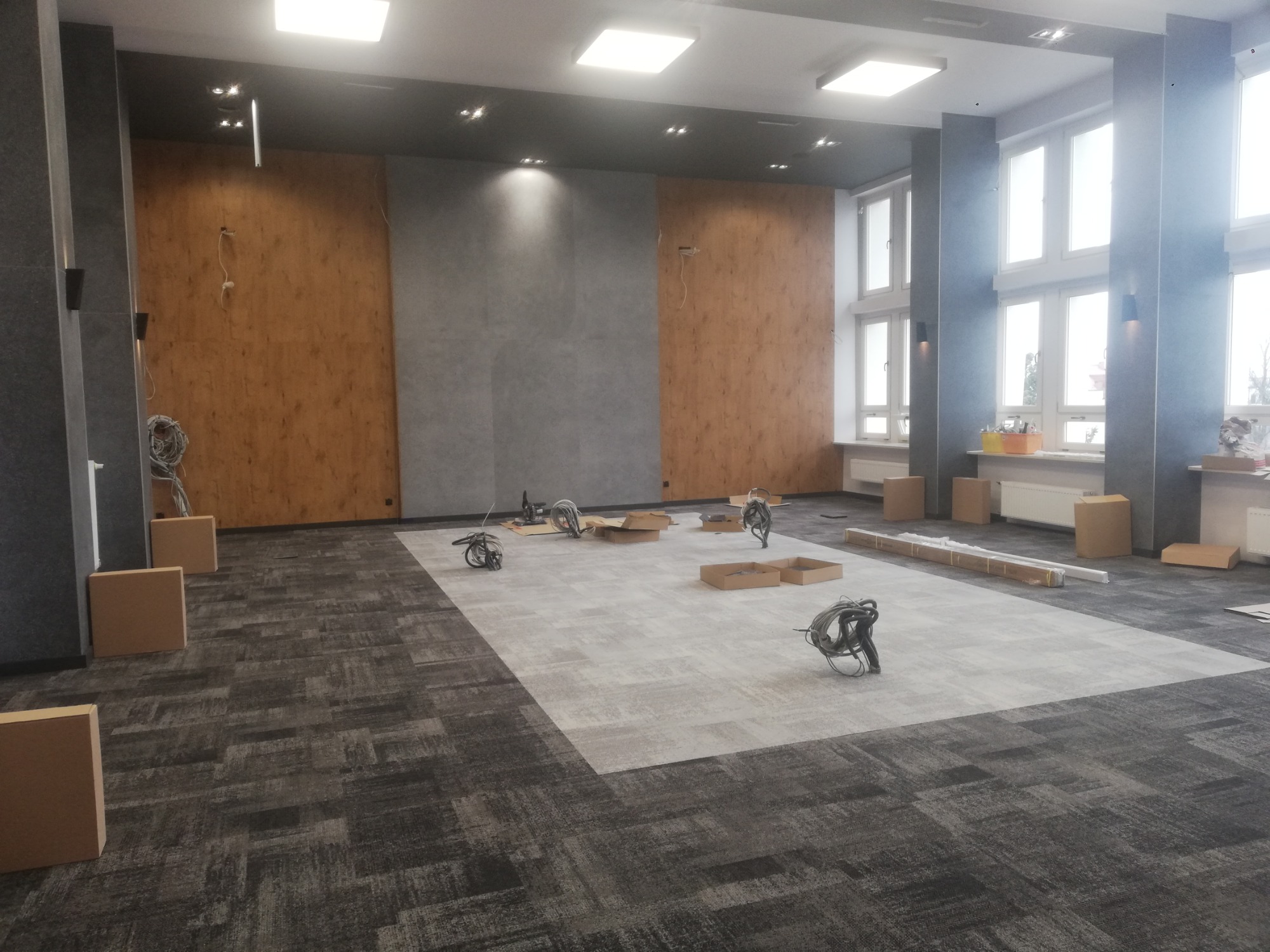 Układanie wzorzystej wykładziny dywanowej w dużym pomieszczeniu biurowym, widoczne narzędzia i kartony, ściany częściowo wykończone panelami drewnopodobnymi i szarymi płytami.