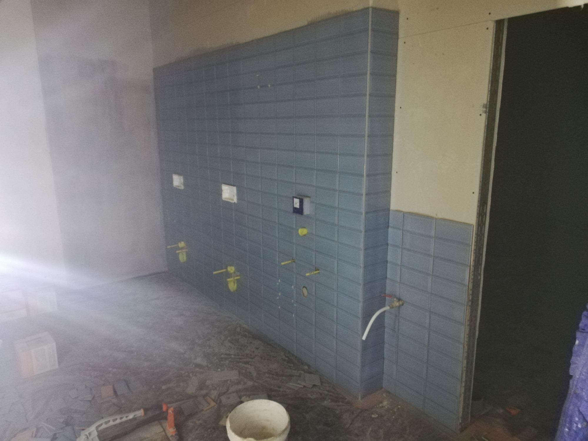 Niedokończona łazienka z niebieskimi płytkami ułożonymi na ścianie, widoczne wystające rury i otwory na instalacje, na podłodze resztki płytek i narzędzia, kubeł na zaprawę.