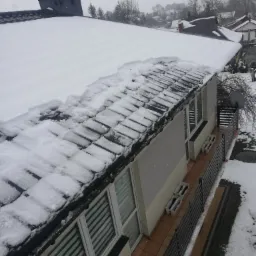 Śnieżny dach domu z czarną dachówką, częściowo odśnieżony, z widocznymi oknami z żaluzjami i balkonem z czarną balustradą w zimowej scenerii.
