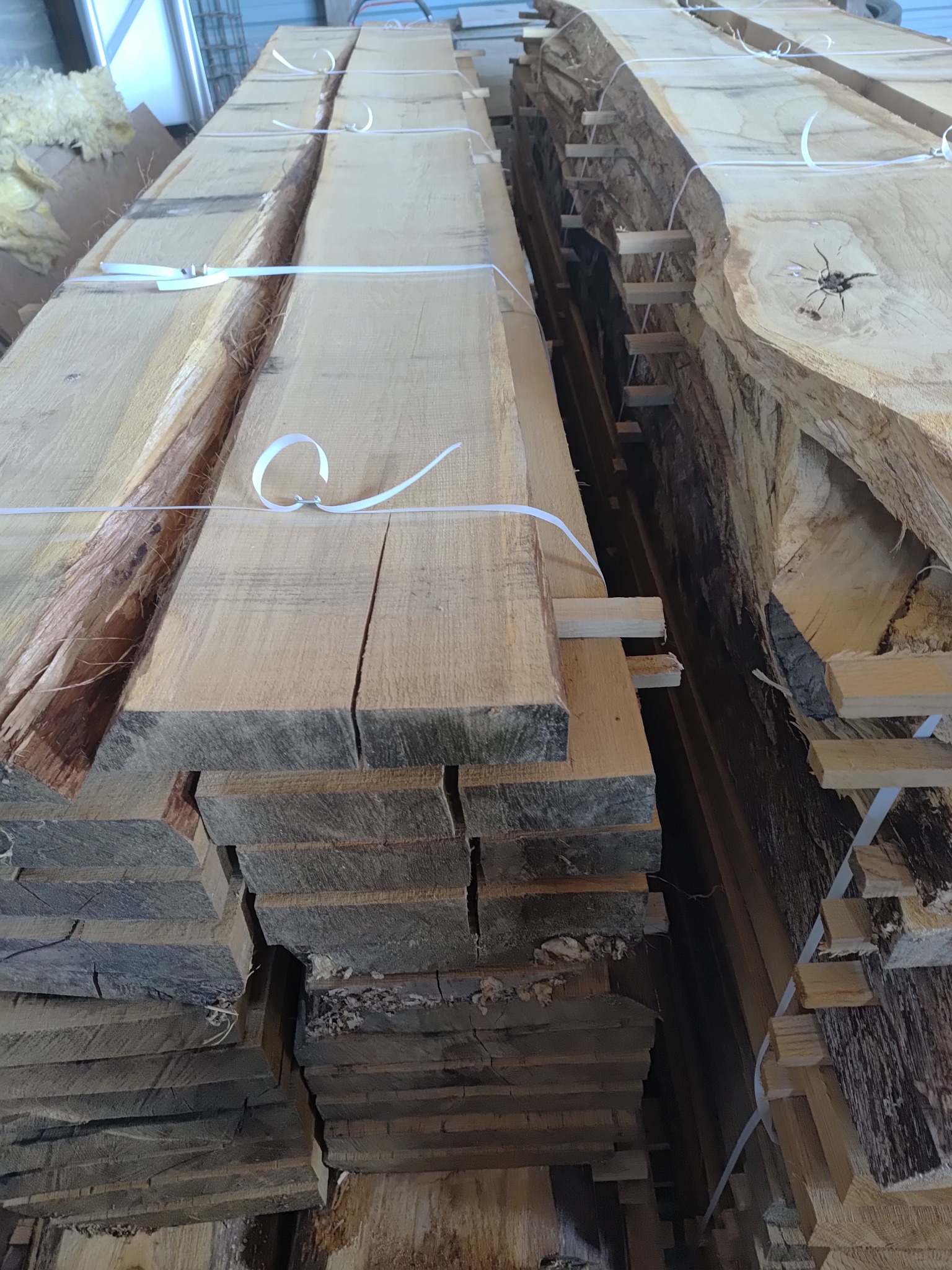 Stosy świeżo przetartego drewna, zabezpieczone białymi taśmami, ukazane w perspektywie od dołu ku górze. Widoczne naturalne krawędzie desek i jasne drewno.