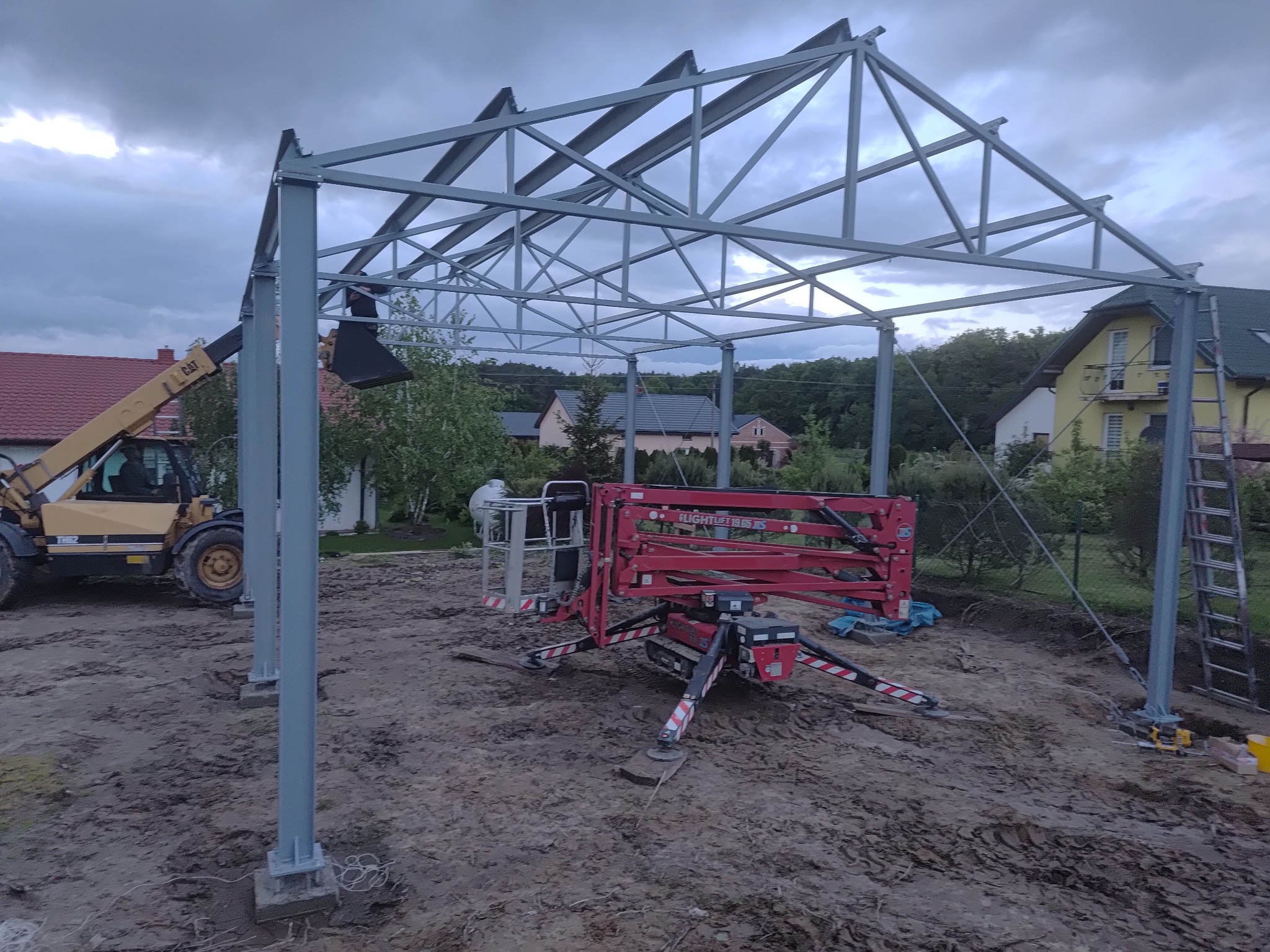 Montaż stalowej konstrukcji z użyciem podnośnika i ładowarki teleskopowej na placu budowy. Widoczne słupy, kratownica dachu, maszyny budowlane i domy w tle.