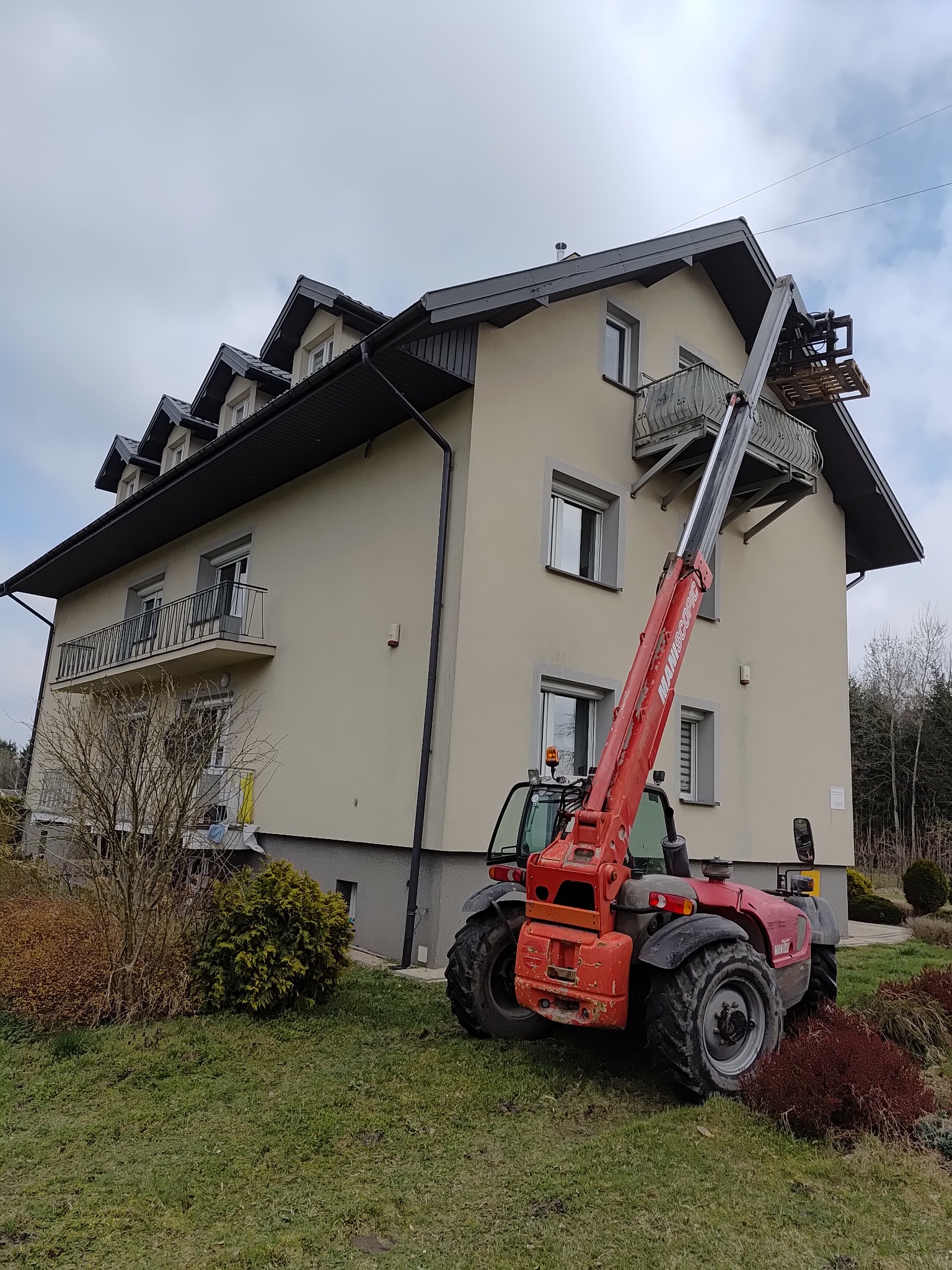 Czerwony podnośnik teleskopowy Manitou z paletą przy balkonie dwupiętrowego domu z szarym dachem i rynnami, na tle zieleni i pochmurnego nieba.