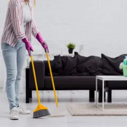 Kobieta w różowych rękawicach zamiata podłogę w pokoju, obok stoi wiadro ze środkami czystości, w tle ciemna kanapa.