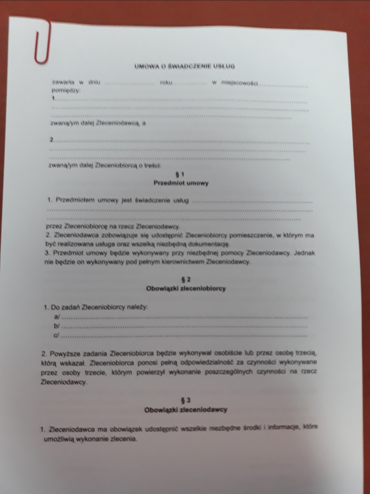 Fragment umowy o świadczenie usług, z widocznym nagłówkiem, paragrafami dotyczącymi przedmiotu umowy, obowiązków zleceniobiorcy i zleceniodawcy, przypiętej czerwoną spinaczem.