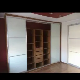 Wnętrze z drewnianą szafą wnękową z przesuwnymi drzwiami z mlecznego szkła, drewniane półki i szuflady wewnątrz, widoczny fragment podłogi i sufitu z oświetleniem.