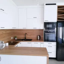 Nowoczesna kuchnia z białymi szafkami, drewnianym blatem i czarną armaturą, uzupełniona o wbudowane sprzęty AGD i regał na wino.