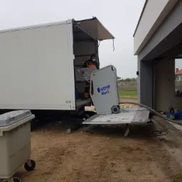 Pracownik w masce wyładowuje z ciężarówki maszynę do wdmuchiwania izolacji Geko Karl Isy, w tle dom z garażem.