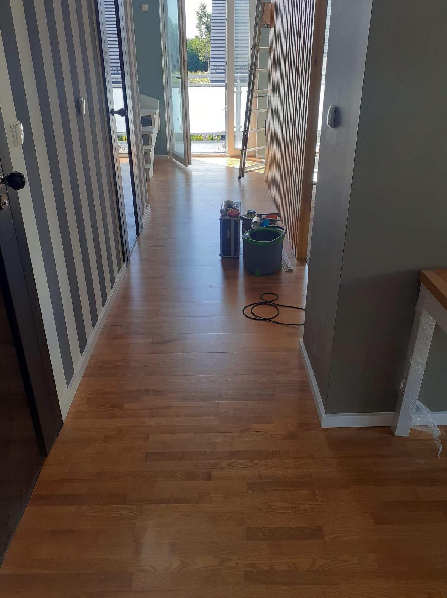 Korytarz w trakcie prac wykończeniowych: świeżo położona drewniana podłoga, ściana w biało-szare pasy, widoczne narzędzia i drabina w tle.