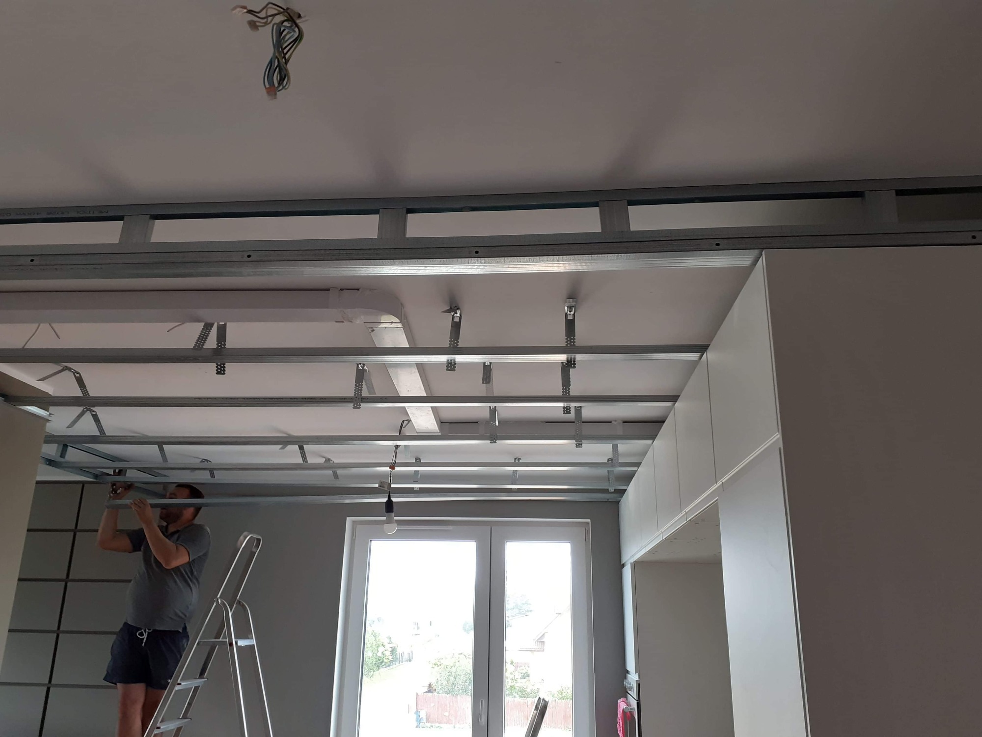Montera na drabinie mocuje metalowy stelaż pod sufitem podwieszanym, widoczne przewody elektryczne, okno i zabudowa kuchenna.