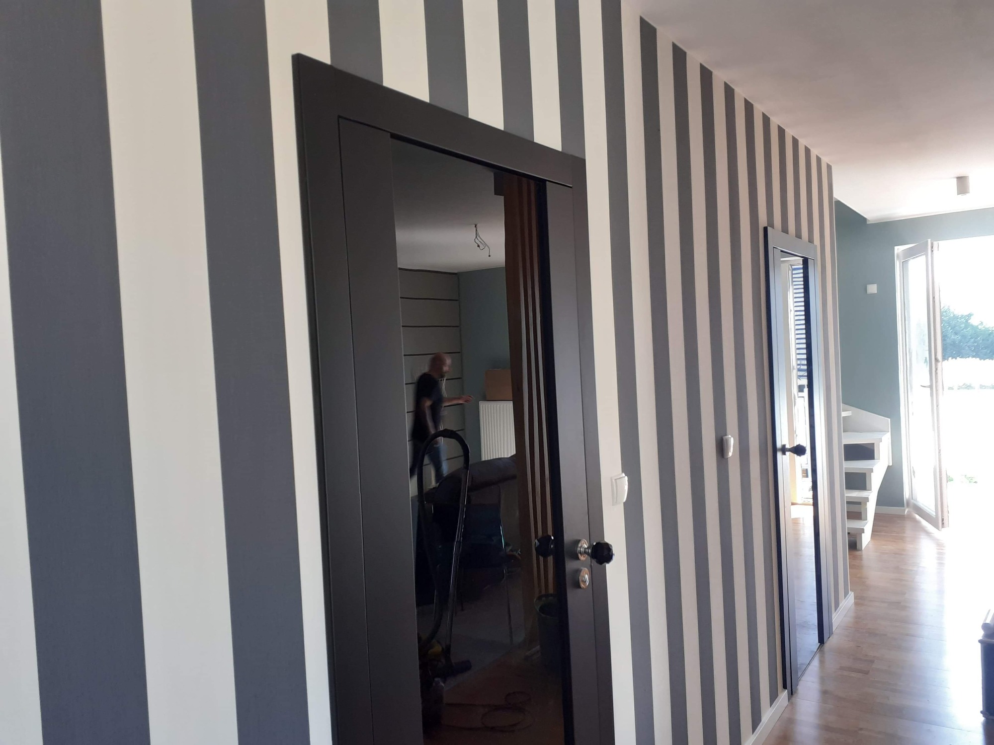 Ciemna, nowoczesna ościeżnica z zamontowanymi drzwiami na tle ściany w szaro-białe pasy. W lustrzanym odbiciu drzwi widoczny fragment wnętrza z elementami wyposażenia i osobą w tle.