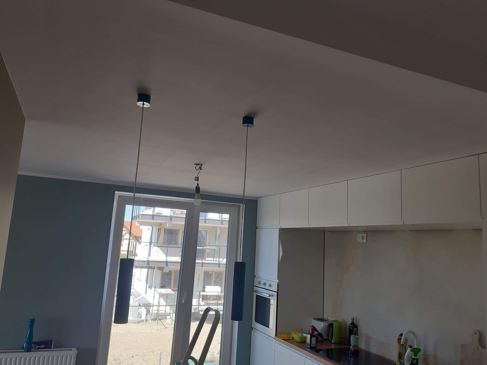 Wnętrze w trakcie odświeżania: widoczne niedomalowane ściany, drabina oparta o okno, wiszące lampy z niebieskimi akcentami i białe szafki kuchenne.