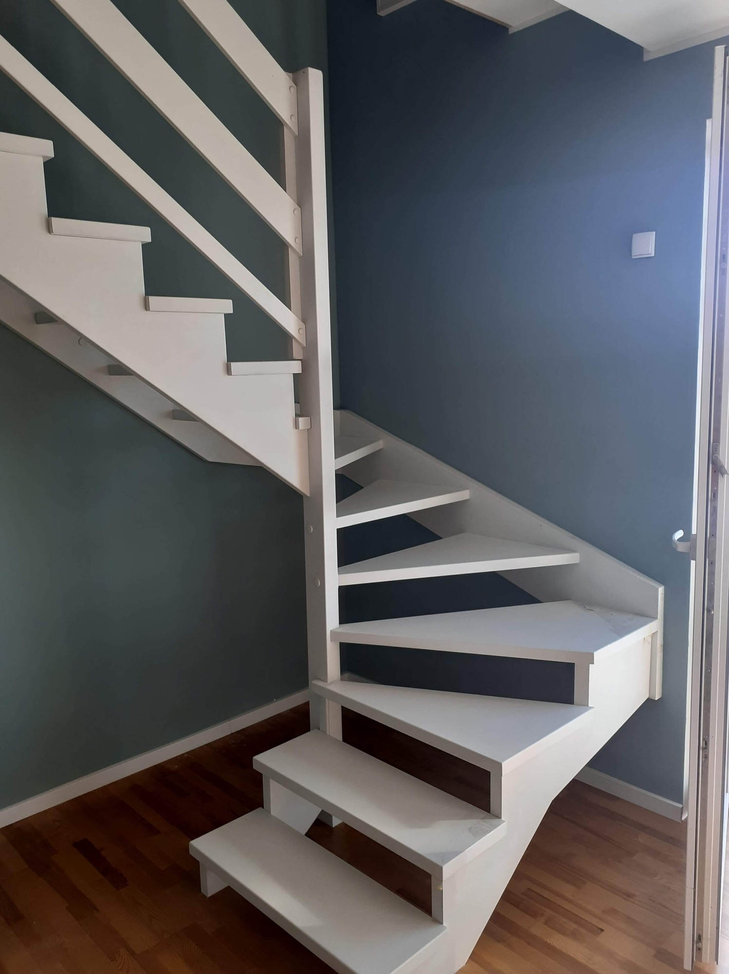 Białe drewniane schody spiralne z balustradą, prowadzące w górę, na tle ściany pomalowanej na ciemny odcień niebieskiego, z widoczną podłogą z paneli drewnianych.