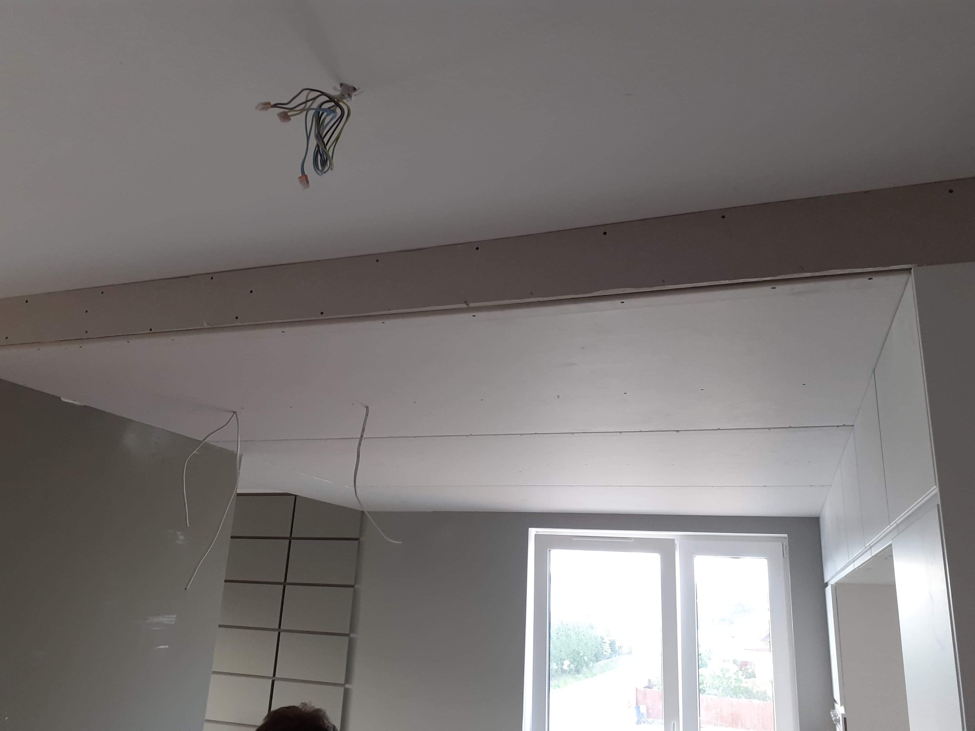 Montaż sufitu podwieszanego z widocznymi przewodami elektrycznymi i profilem mocującym, na tle okna i częściowo wykończonej ściany.