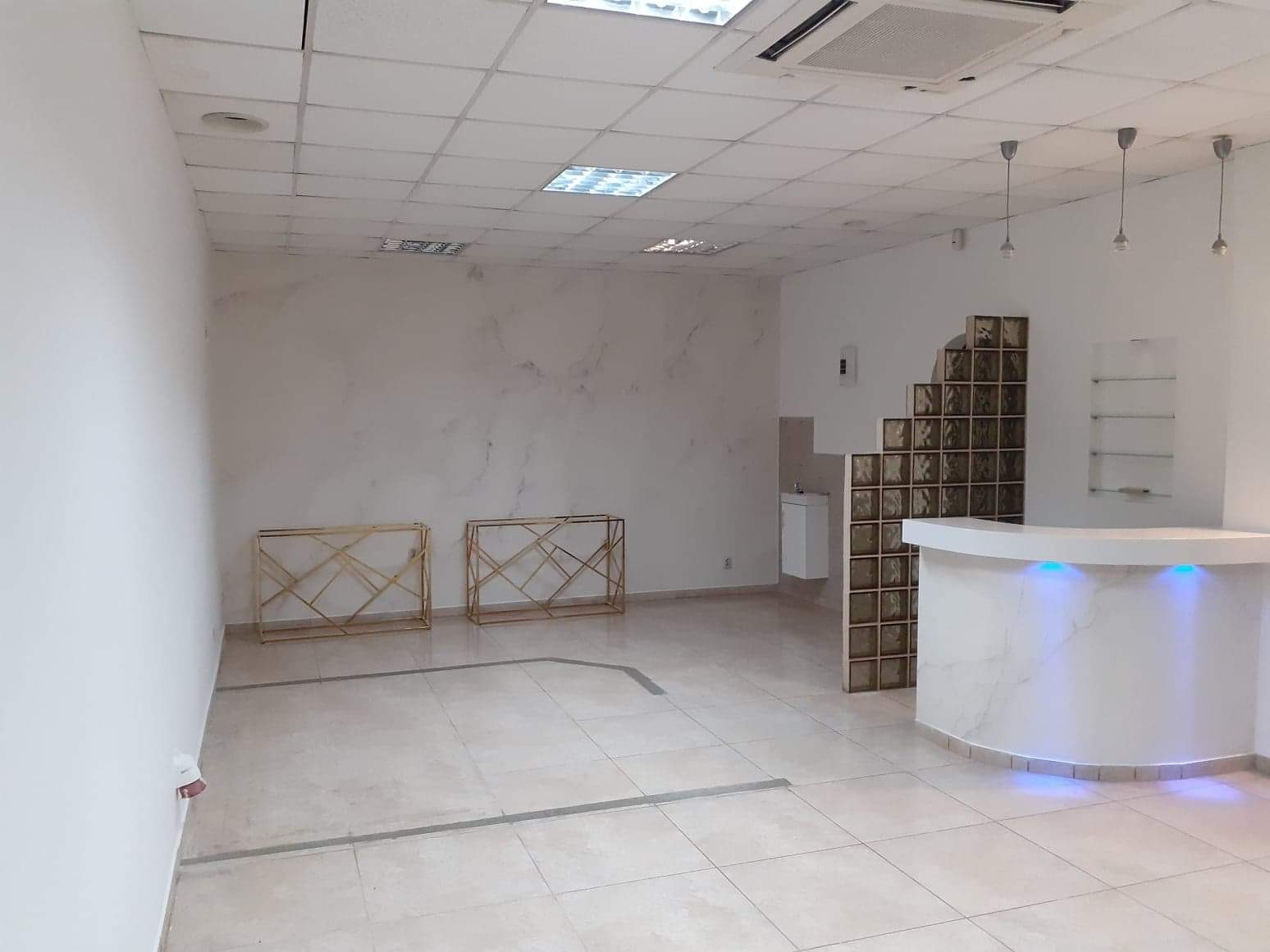 Wnętrze lokalu usługowego w trakcie przygotowania: białe ściany z marmurkowym wzorem, podłoga wyłożona jasnymi płytkami, podświetlany ladą barowa z elementami szklanych pustaków, dwie metalowe...