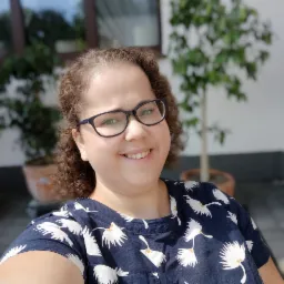 Portret uśmiechniętej kobiety w okularach i bluzce z motywem białych ptaków na tle białej ściany i zielonych roślin w donicach.