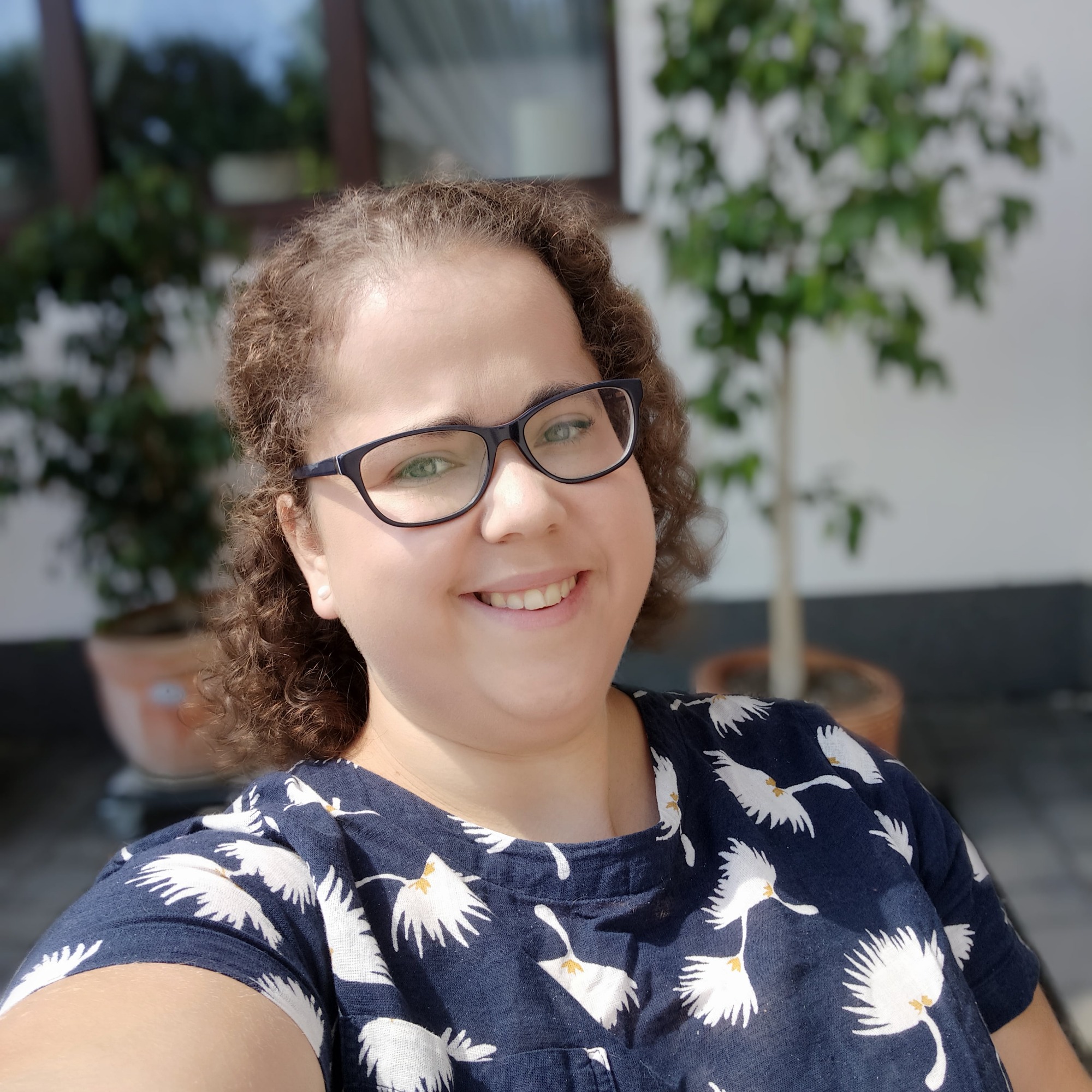 Portret uśmiechniętej kobiety w okularach i bluzce z motywem białych ptaków na tle białej ściany i zielonych roślin w donicach.