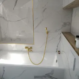 Nowoczesna łazienka z białą wanną obudowaną płytkami imitującymi marmur, armaturą w kolorze złotym i półką z drewnianym blatem.