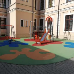 Kolorowy plac zabaw z huśtawkami, zjeżdżalnią i sprężynową zabawką na tle beżowego budynku z ciemnymi oknami.