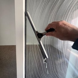 PARIX - Mycie okna z użyciem ściągaczki, widoczne ślady piany. Ręka w trakcie pracy, okno w białej ramie, fragment ściany z teksturą w tle.